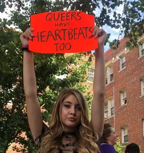 A young woman holds a sign that says "Queers have heartbeats, too" in all capital letters. 