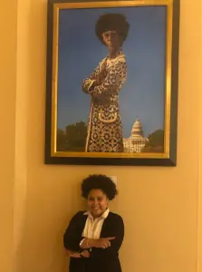 VCI Janiah poses below a Shirley Chisholm portrait with the same crossed arm pose as Shirley Chisholm
