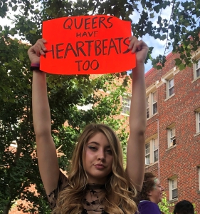 A young woman holds a sign that says "Queers have heartbeats, too" in all capital letters. 