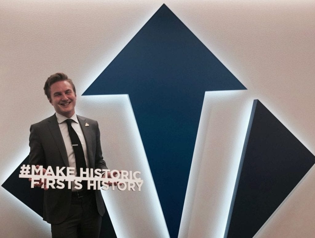 Brett Ries poses in front of Victory sign with "make historic firsts history" sign